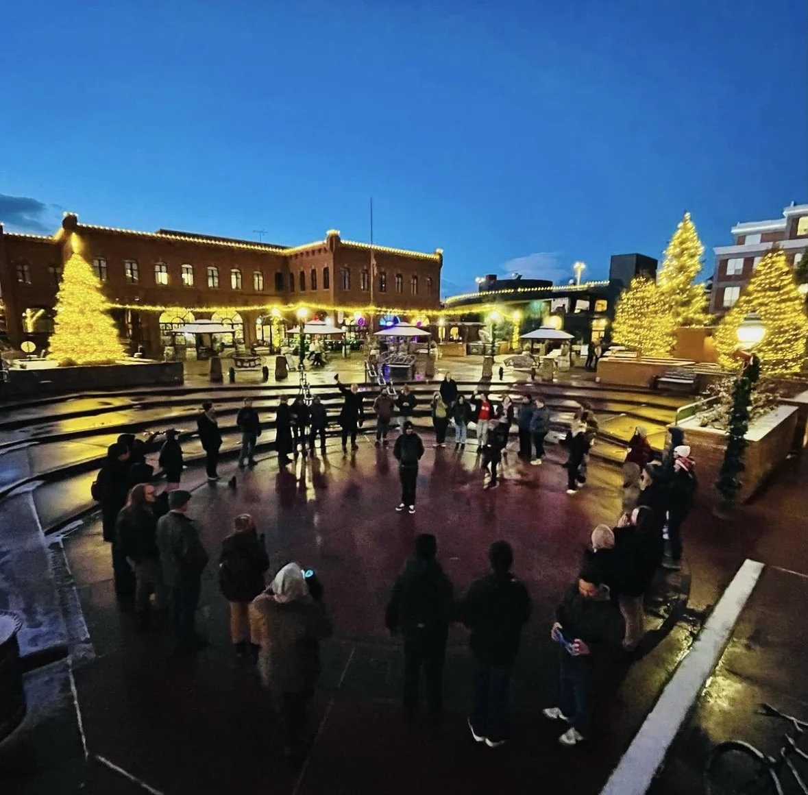 Unsilent Night participants gathered in a circle in a plaza decorated with lit Christmas trees at dusk