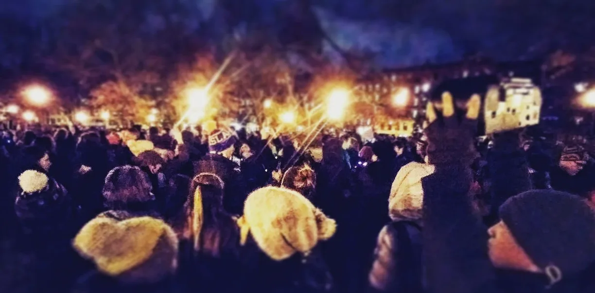 Crowd of Unsilent Night participants gathered at dusk in a park, street lights glowing through bare trees, one person holding a boombox overhead