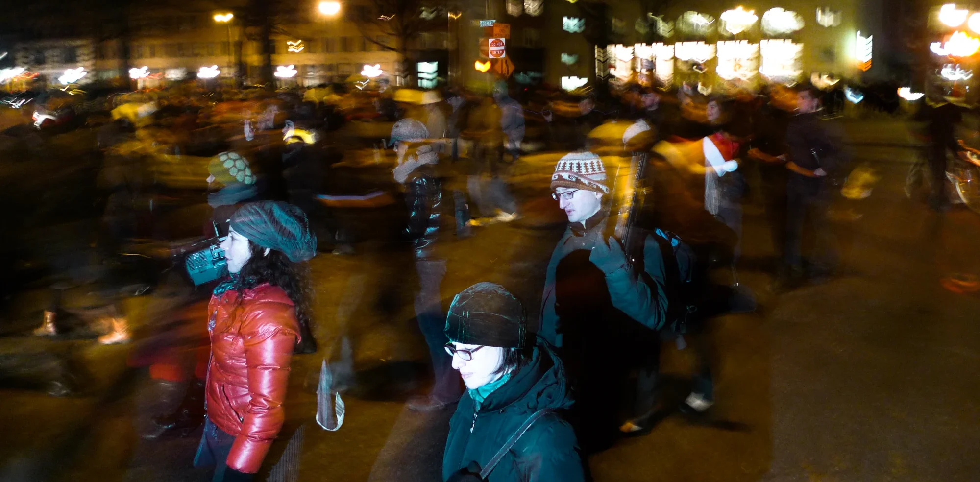 Unsilent Night participants walking at night in a long-exposure blur: a woman in a red jacket carrying a boombox beside two others in winter hats, city lights behind them